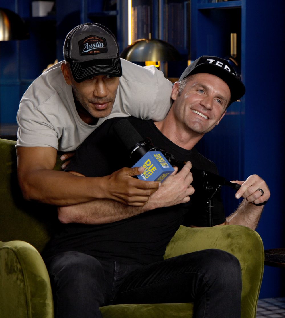 Bill Perkins, in his grey Austin cap, hugging Austin Allison from behind his chair in the Die With Zero studio, both laughing
